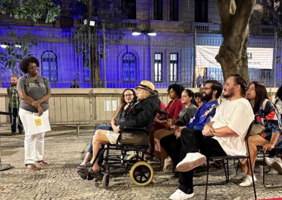 Em uma praça à noite, um grupo de pessoas participa de um evento cultural ao ar livre. À esquerda, uma mulher em pé fala ao público segurando papéis. À frente dela, um homem idoso em cadeira de rodas usa chapéu claro e segura um microfone. Outras pessoas estão sentadas em cadeiras, observando atentamente. Ao fundo, grades, árvores e um prédio iluminado com luz azul compõem o cenário urbano.