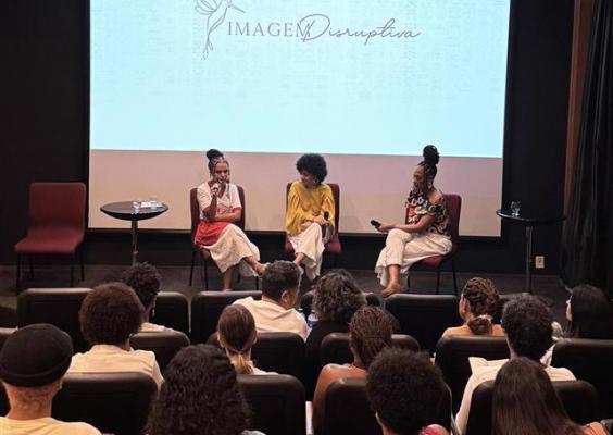 Auditório com público sentado assistindo a uma roda de conversa no palco. Três mulheres negras estão sentadas em cadeiras, segurando microfones e dialogando diante de uma grande tela de projeção. A tela exibe o nome “Imagem Disruptiva” com o desenho de um pássaro. Pequenas mesas laterais com copos de água completam o cenário. O ambiente é acolhedor e voltado para debate cultural e artístico.