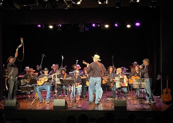 Palco de teatro iluminado recebe um grande grupo de músicos durante apresentação ao vivo. Homens e mulheres, muitos usando chapéus, tocam violões, acordeão e outros instrumentos sentados em semicírculo. No centro, um homem de costas conduz a apresentação com violão. À esquerda, outro participante ergue um berrante. O fundo escuro destaca as luzes do palco e o clima de música regional e celebração cultural.