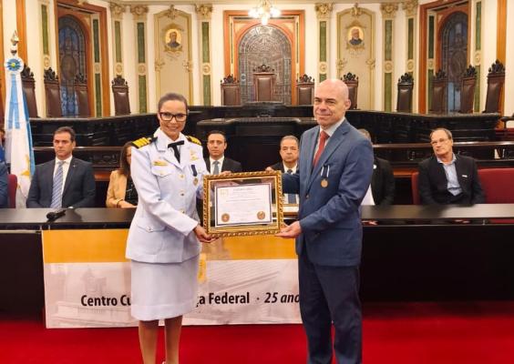 Fotografia de uma cerimônia oficial realizada em um salão histórico de arquitetura clássica, com teto ornamentado, vitrais e cadeiras de madeira escura ao fundo. No centro, uma mulher de uniforme branco da Marinha Mercante e um homem de terno azul posam lado a lado, segurando um diploma emoldurado. Ambos sorriem para a câmera. Atrás deles, autoridades e convidados estão sentados em uma bancada elevada. No piso, um tapete vermelho cobre toda a área. Uma faixa alusiva aos 25 anos do Centro Cultural Justiça Fe