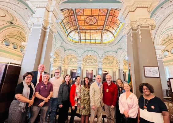 Fotografia de grupo em um amplo salão histórico de arquitetura ornamentada. Doze pessoas posam lado a lado, sorrindo para a câmera, em frente a colunas altas e arcos decorados em tons claros. Ao fundo, destaca-se um grande vitral colorido no teto, que ilumina o ambiente com luz suave. Os participantes usam roupas variadas, em tons neutros e vermelhos. À direita, vê-se parcialmente uma bandeira do Brasil. A imagem transmite um clima institucional e de confraternização.