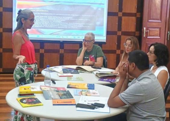 Atividade formativa em sala. À esquerda, uma mulher de cabelos grisalhos presos, óculos e blusa rosa sem mangas fala ao grupo, gesticulando com uma das mãos. Ao redor de uma mesa redonda, quatro participantes escutam e consultam livros e materiais impressos. Sobre a mesa há diversos livros, cadernos, canetas, um celular e uma garrafa de água. Ao fundo, uma tela de projeção exibe uma apresentação de slides. O ambiente tem paredes revestidas em madeira e atmosfera de oficina literária ou encontro de leitura.
