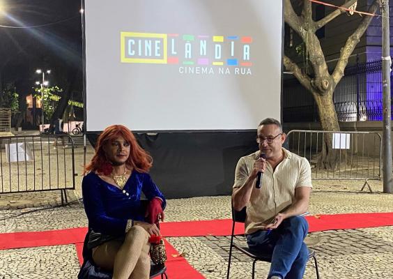 Willa Queer e Celso André participam de conversa ao ar livre durante o projeto Cinelândia: Cinema na Rua. Sentados diante de um telão de projeção, Willa Queer veste figurino azul com peruca ruiva e acessórios dourados, enquanto Celso André segura um microfone e fala ao público. Ao fundo, o logotipo do projeto é projetado na tela, e o espaço aberto com árvores e calçamento de pedras compõe o ambiente da atividade.