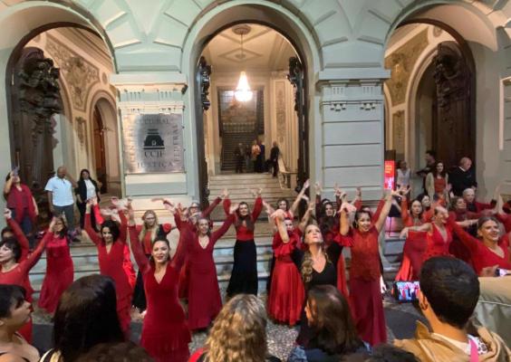 Integrantes do grupo Gesto realizam apresentação em frente ao Centro Cultural Justiça Federal. Vestindo figurinos em diferentes tons de vermelho, dezenas de artistas ocupam a escadaria e a entrada do edifício com os braços erguidos em uma coreografia coletiva. Ao fundo, destacam-se a fachada histórica do CCJF, seus grandes arcos e o hall iluminado, enquanto o público acompanha a apresentação da calçada.