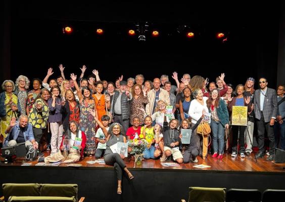 Fotografia de grupo no Teatro do Centro Cultural Justiça Federal. Cerca de cinquenta pessoas, entre adultos, idosos e crianças, posam reunidas sobre o palco, muitas delas sorrindo e acenando para a câmera. Alguns participantes seguram livros e obras de arte, enquanto outros exibem flores. Ao fundo, o palco escuro e a iluminação cênica destacam o grupo em clima de celebração e encerramento de atividade cultural.