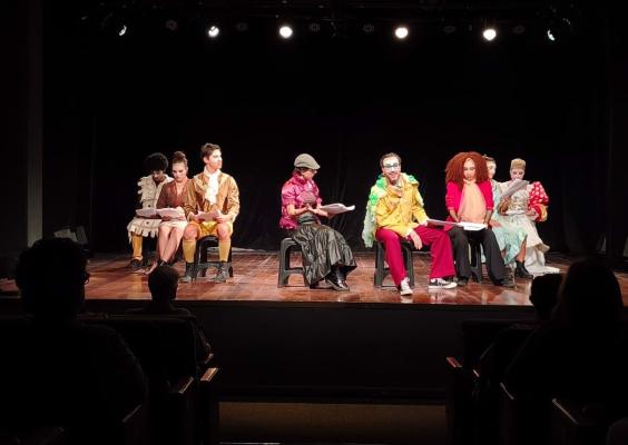 Cena teatral no palco do Teatro do Centro Cultural Justiça Federal. Oito artistas, usando figurinos variados e coloridos, estão sentados em cadeiras e seguram roteiros ou folhas de texto durante a apresentação. Ao centro, um personagem com maquiagem cênica e roupas amarelas e vermelhas contrasta com os demais, enquanto a plateia aparece em silhueta no primeiro plano, reforçando a perspectiva de quem assiste ao espetáculo.
