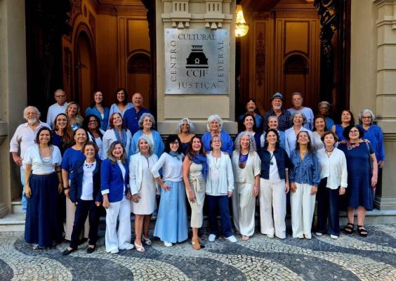 O Coral da Tarde reúne cerca de trinta e cinco integrantes para uma fotografia em frente à entrada principal do Centro Cultural Justiça Federal. Vestindo roupas em tons de azul, branco e bege claro, os participantes sorriem para a câmera diante da fachada histórica do edifício, onde se destaca a placa com o nome do CCJF e o hall de entrada iluminado.