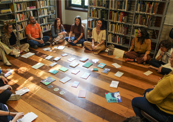 Participantes da oficina de investigação criativa sentam-se em círculo no chão da biblioteca do Centro Cultural Justiça Federal. No centro da roda, diversos livros estão espalhados sobre o piso de madeira, formando um espaço de leitura e compartilhamento. Ao fundo, estantes repletas de livros e a luz natural que entra pela janela criam um ambiente acolhedor de escuta, reflexão e criação.