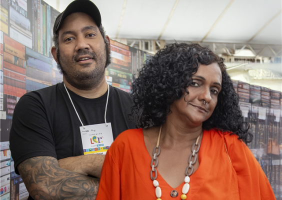 Retrato do diretor Márcio Coutinho e da pesquisadora e produtora Rachel Silva, do documentário Dos Armários para as Ruas. À esquerda, Márcio Coutinho veste camiseta preta e boné, mantém os braços cruzados e usa crachá de identificação. À direita, Rachel Silva veste blusa laranja e colar de contas, olhando para a câmera com expressão serena. Ao fundo, um painel formado por lombadas de livros reforça o ambiente cultural.