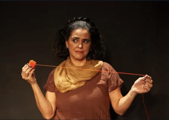 Cena do espetáculo Filipa, no Teatro do Centro Cultural Justiça Federal. A atriz Waleska Arêas aparece diante de fundo escuro segurando um carretel de linha vermelha. Com os braços abertos, ela mantém o fio esticado entre as mãos enquanto dirige o olhar para um dos lados, com expressão reflexiva. Veste blusa marrom e lenço em tom bege, e a iluminação concentra a atenção na atriz e na linha vermelha, elemento central da composição.