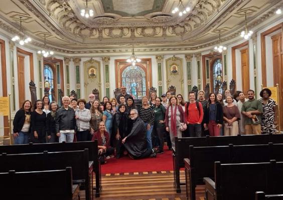 Fotografia de grupo na Sala de Sessões do Centro Cultural Justiça Federal. Cerca de trinta participantes posam sorrindo para a câmera, reunidos em frente às cadeiras da mesa diretora. Ao fundo, destacam-se o vitral central, os retratos históricos, os ornamentos do teto e a arquitetura neoclássica do espaço, um dos ambientes mais emblemáticos do CCJF.