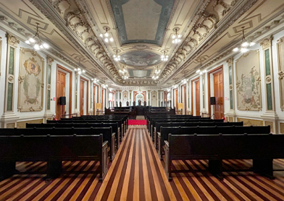 Imagem da Sala de Sessões do Centro Cultural Justiça Federal (CCJF), com vista frontal para o espaço. A foto mostra fileiras de bancos de madeira escura alinhadas em direção ao palco ao fundo, onde se vê uma mesa cerimonial e um vitral colorido. O piso tem listras em tons claros e escuros, o teto é ricamente decorado com afrescos e molduras em relevo, e as paredes exibem painéis artísticos com detalhes dourados. A iluminação clara ressalta a elegância e imponência do ambiente.
