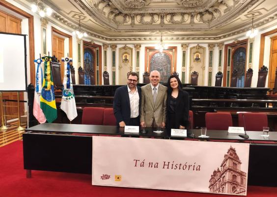 Foto de um salão histórico, ricamente ornamentado, com teto e paredes decorados em estilo clássico. Ao centro, três pessoas posam atrás de uma longa mesa, diante de cadeiras de madeira escura. À esquerda, há um púlpito e três bandeiras, incluindo a do Brasil. Na frente da mesa, um painel traz a frase “Tá na História” e a imagem de um prédio histórico.