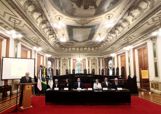 Foto de cerimônia na Sala de Sessões do CCJF, com teto ricamente ornamentado e lustres. Ao fundo, vitrais e cadeiras de madeira. À frente, uma mesa reúne sete pessoas sentadas, entre elas uma mulher uniformizada, e há bandeiras ao lado. À esquerda, um homem de terno fala ao microfone, diante de um telão. O piso e o tapete são vermelhos.