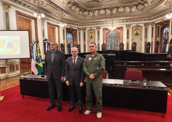 Foto de homenagem na Sala de Sessões do CCJF. Em primeiro plano, três homens posam lado a lado diante de uma longa mesa: dois usam ternos escuros e gravatas, e o terceiro veste uniforme verde de aviador, com insígnias e distintivos. Ao fundo, veem-se as cadeiras da sala, vitrais, retratos, bandeiras e o teto ricamente ornamentado. O ambiente tem carpete vermelho.