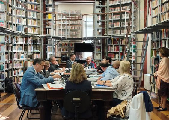 Grupo de cerca de dez pessoas participa de uma reunião em torno de uma mesa retangular em uma biblioteca. Os participantes usam roupas sociais, consultam documentos e conversam. Ao fundo, estantes altas repletas de livros ocupam toda a parede, com passarelas e escadas metálicas para acesso aos andares superiores. Uma tela exibe a imagem de uma pessoa em videoconferência. À direita, uma mulher permanece em pé observando a reunião.