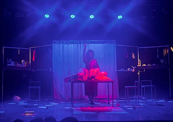 Cena da peça Se Eu Fosse Eu, Clarices. Sob iluminação azul e avermelhada, três atrizes que representam uma mesma mulher ocupam diferentes planos do palco. Ao centro, uma delas está deitada sobre uma mesa, enquanto outra se posiciona atrás dela e uma terceira aparece mais ao fundo, parcialmente encoberta por uma cortina translúcida. Nas laterais do cenário, mesas, bancos e pequenos objetos compõem o ambiente, envolvido por uma atmosfera intimista e onírica.