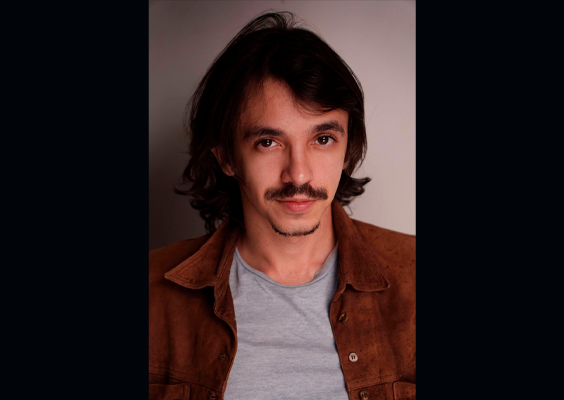 Retrato de Eduardo Segura, diretor de produção da Mostra Vem Ver. Ele aparece de frente para a câmera, com cabelos escuros na altura do pescoço e bigode, vestindo camiseta cinza e camisa marrom aberta. O fundo neutro e a iluminação suave destacam seu rosto.