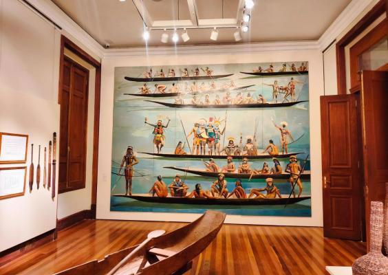 Vista de uma das galerias do CCJF dedicada às obras de Carybé. Um grande painel ocupa toda a parede ao fundo, retratando povos originários em diferentes cenas, com numerosas figuras e canoas sobre fundo azul-claro. No centro da sala há uma canoa de madeira e, na parede lateral, estão expostos dois quadros e diferentes artefatos. O conjunto destaca a presença e a importância dos povos originários para o Brasil.