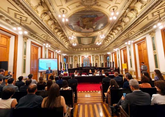 Na histórica Sala de Sessões do CCJF, autoridades participam de cerimônia. Diante da mesa da cerimônia, homenageados ocupam as cadeiras da sala, enquanto o diretor-geral do CCJF fala ao púlpito. O ambiente é marcado pelo piso de madeira, grandes portas, vitrais e pelo teto ornamentado com pinturas e detalhes dourados.