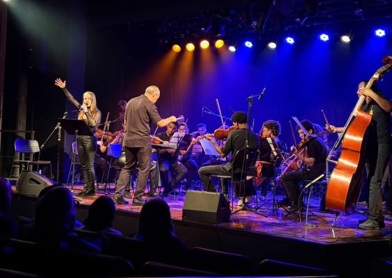 Apresentação do Rock in Concert no palco do CCJF. Sob iluminação azul e amarela, uma orquestra executa clássicos do rock, com músicos tocando instrumentos de cordas, entre eles violinos, violoncelos e contrabaixo. À frente do conjunto, uma cantora se apresenta ao microfone enquanto o maestro rege os músicos. Parte do público aparece em primeiro plano, acompanhando o concerto.