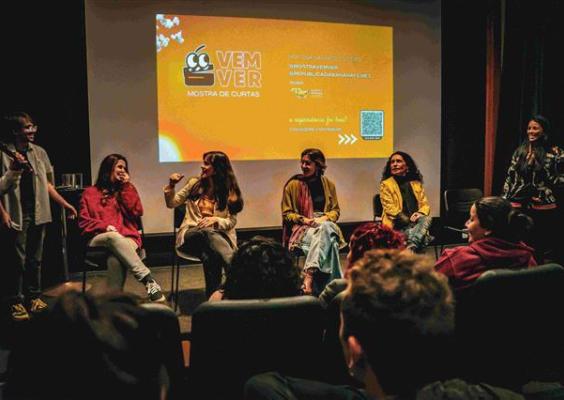 Encontro da Vem Ver – Mostra contínua de curtas no CCJF. Após a exibição dos filmes, realizadores e convidados conversam com o público, reunidos em cadeiras diante da tela de cinema, onde é projetada a identidade visual da mostra. Uma das participantes fala ao microfone enquanto os demais integrantes da roda a observam. Em primeiro plano, parte da plateia acompanha o bate-papo.