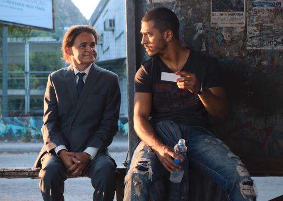 Cena do filme Papagaios. Em um ponto de ônibus, dois homens estão sentados lado a lado. À esquerda, um homem de baixa estatura veste terno cinza, camisa branca e gravata, mantendo as mãos apoiadas sobre o colo enquanto sorri discretamente para o homem ao lado. À direita, um homem de camiseta escura e calça jeans rasgada segura um pequeno cartão branco e uma garrafa de água, retribuindo o olhar do companheiro. Ao fundo, a paisagem urbana é iluminada pela luz dourada do fim da tarde.