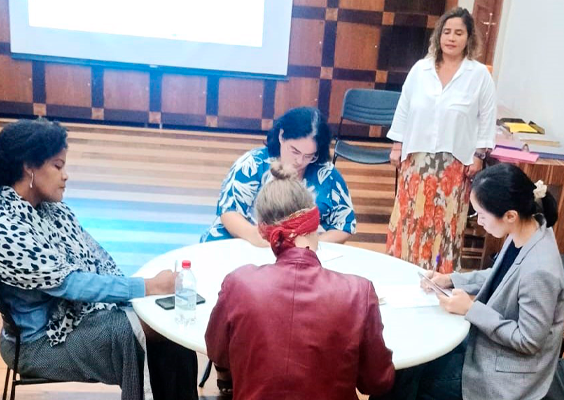 Participantes da Oficina de Literatura dedicada à obra de João Cabral de Melo Neto realizam uma atividade em grupo no CCJF. Quatro pessoas estão sentadas ao redor de uma mesa, lendo e fazendo anotações em papéis, enquanto uma mulher em pé acompanha o trabalho. Ao fundo, uma tela de projeção e cadeiras compõem o ambiente da oficina.