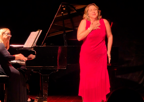 Recital de ópera no Teatro do CCJF. No palco, uma cantora de vestido longo vermelho interpreta uma obra com uma das mãos sobre o peito, enquanto, à esquerda, uma pianista de vestido preto toca um piano de cauda. A iluminação destaca as duas artistas contra o fundo escuro do palco, e parte da plateia aparece em silhueta em primeiro plano.