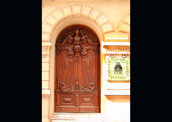 Porta principal de entrada do Centro Cultural Justiça Federal (CCJF), em madeira escura, com entalhes ornamentais e arco na parte superior. Ao lado, uma placa identifica o Centro Cultural Justiça Federal junto ao símbolo da Justiça. A entrada é emoldurada pela fachada histórica em tom claro.