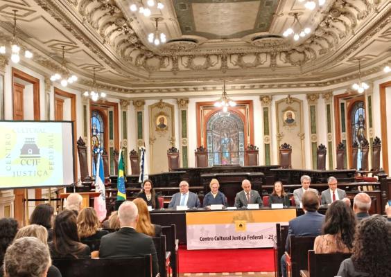 Em um salão histórico e ornamentado, seis pessoas estão sentadas à mesa de um evento, diante de uma plateia. Ao fundo, há vitrais, esculturas, pinturas, bandeiras do Brasil e outras, além de detalhes dourados no teto e nas paredes. Um telão à esquerda exibe o logotipo do Centro Cultural Justiça Federal.