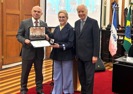 Três pessoas posam em um salão histórico. A mulher ao centro segura, com o homem à esquerda, uma placa de homenagem aberta, com certificado e medalha. O homem à direita sorri ao lado deles. Ao fundo, há uma tela de projeção, bandeiras do Brasil e de outros países, mobiliário de madeira escura e detalhes ornamentais. O piso é de madeira e há um tapete vermelho.