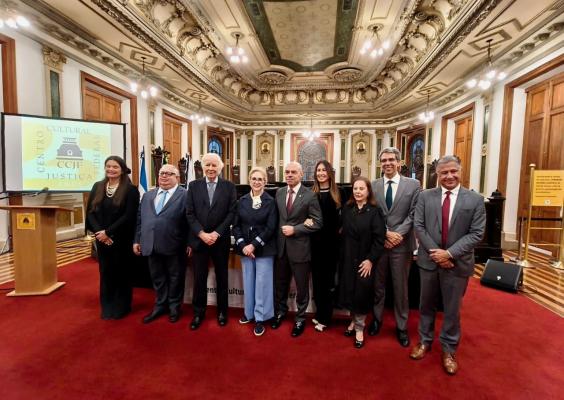 Oito pessoas posam lado a lado em um salão histórico, sobre um tapete vermelho. Ao fundo, destacam-se um teto ricamente ornamentado, vitrais, painéis decorativos, portas de madeira, bandeiras e o logotipo do Centro Cultural Justiça Federal em uma tela. As pessoas usam roupas sociais, e uma delas, ao centro, veste blazer escuro e calça azul-clara.