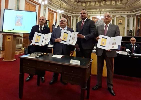 Foto de quatro homens em um salão histórico, com decoração ornamentada, lustres e carpete vermelho. Eles usam ternos e seguram pastas abertas com imagens de selos comemorativos. Ao fundo, há uma tela de projeção e, à direita, um homem sentado atrás de uma mesa. Em primeiro plano, uma mesa de madeira exibe documentos e uma placa com a marca dos Correios.