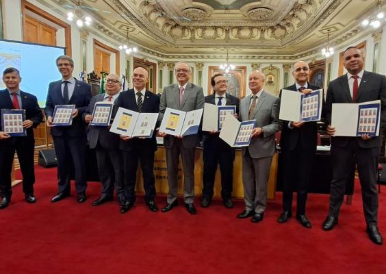Foto de grupo em um salão histórico e ornamentado. Oito homens, vestidos de terno e gravata, estão lado a lado sobre um carpete vermelho. Alguns seguram folhas ou pastas com imagens de selos comemorativos. Ao fundo, há painéis de madeira, portas, pinturas e um teto ricamente decorado. Todos posam voltados para a câmera.