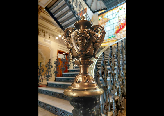 Detalhe da escadaria principal do CCJF, com ornamento metálico dourado em primeiro plano, integrado ao corrimão de ferro trabalhado. Ao fundo, aparecem os degraus da escada e parte dos vitrais do edifício histórico.