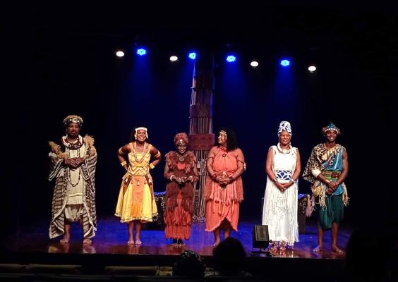 No palco do Teatro do CCJF, seis artistas estão alinhados diante do público, usando figurinos coloridos de inspiração africana, com vestidos, túnicas, estampas e adereços de cabeça. Ao fundo, uma estrutura cenográfica vertical ocupa o centro do palco, iluminado predominantemente por luz azul.