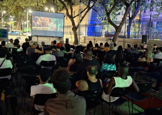 Público assiste à exibição do filme Papagaios no Cinelândia Cinema na Rua. À noite, dezenas de pessoas estão sentadas em cadeiras diante de uma grande tela instalada ao ar livre. Árvores, grades e a iluminação azulada do entorno compõem o espaço da sessão.