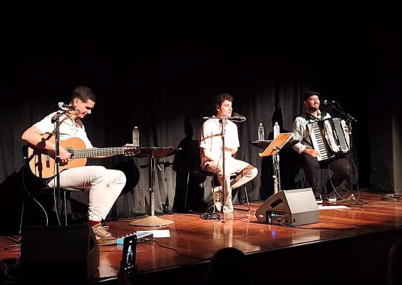 Trio Três em Travessia se apresenta no palco do Teatro do CCJF. À esquerda, um músico toca violão; ao centro, outro integrante está sentado diante de um microfone; e, à direita, o terceiro toca acordeão e canta ao microfone. Partituras, caixas de som e outros equipamentos estão distribuídos pelo palco.