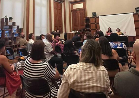 Público participa de uma roda de conversa em uma sala do CCJF. As pessoas estão sentadas em cadeiras voltadas para duas participantes posicionadas à frente do espaço, junto a uma mesa e uma tela de projeção. Ao fundo, portas altas de madeira e estantes com livros compõem o ambiente.