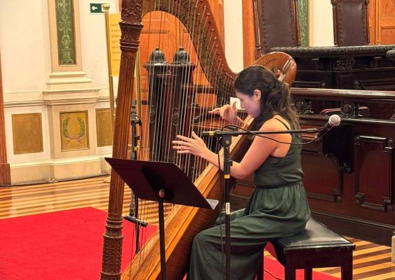 Harpista se apresenta na Sala de Sessões do CCJF. Sentada diante de uma grande harpa de madeira, ela toca o instrumento com as duas mãos, com uma partitura à frente e um microfone posicionado ao lado. O piso de madeira, o tapete vermelho e o mobiliário histórico da sala aparecem ao redor.