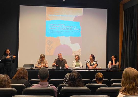Roda de conversa no Cinema do CCJF. Cinco participantes estão sentados à mesa diante do público, com microfones e materiais à frente. À esquerda, uma intérprete de Libras acompanha o encontro em pé. Ao fundo, uma projeção exibe a identidade visual do evento. Na parte inferior da imagem, aparecem participantes da plateia sentados nas poltronas.