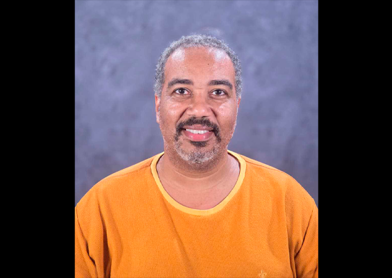 Retrato de Leandro Santanna, sorrindo para a câmera, com camiseta em tom alaranjado, diante de fundo cinza-azulado.