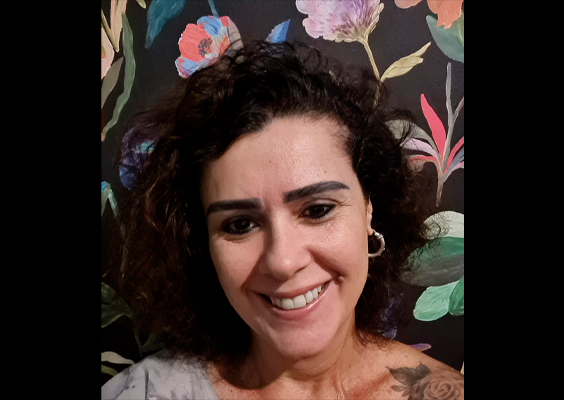 Retrato de Luciana Villar sorrindo para a câmera, com cabelos escuros e cacheados na altura dos ombros. Ela usa brincos de argola e aparece diante de um fundo escuro decorado com flores e folhagens coloridas.