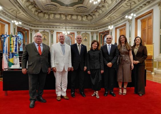 Fotografia de sete pessoas, quatro homens e três mulheres, posando lado a lado em um salão solene, sobre um tapete vermelho. Ao fundo, há cadeiras de madeira escura, vitrais coloridos, bandeiras e paredes com detalhes ornamentais dourados. O teto é ricamente decorado com relevos e lustres. Os participantes vestem trajes formais e sorriem para a foto