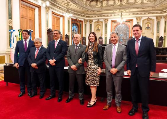 Seis pessoas posam lado a lado em um salão suntuoso, com piso vermelho, paredes ornamentadas e teto ricamente decorado. Há quatro homens de terno à esquerda e à direita, outro homem ao centro e uma mulher entre eles, usando vestido estampado de oncinha e jaqueta preta. Ao fundo, veem-se portas de madeira, vitrais, cadeiras e bandeiras.