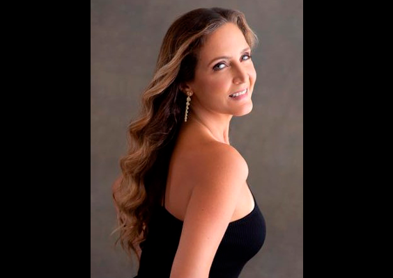 Retrato de Eliane Carvalho, de costas com o rosto virado para a câmera e sorrindo. Veste um vestido preto sem alças; o cabelo é castanho, ondulado e comprido.