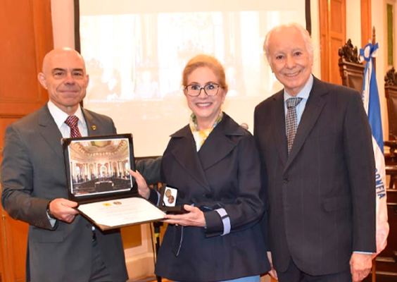 Três pessoas posam para uma foto em um salão histórico. À esquerda, um homem careca de terno cinza segura uma placa comemorativa aberta, com uma fotografia do salão e um certificado. Ao centro, uma mulher de óculos, casaco escuro e calça azul-clara segura a homenagem e sorri. À direita, um homem de cabelos brancos, usando terno escuro e gravata, também sorri. Ao fundo, há portas de madeira, uma tela de projeção e uma bandeira.