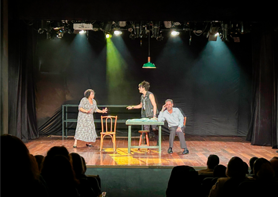 Cena teatral em um palco com iluminação colorida, onde três atores interpretam uma situação ao redor de uma mesa. À esquerda, uma mulher de vestido estampado gesticula; ao centro, um homem de colete sem mangas estende o braço em sua direção; à direita, outro homem está sentado, com expressão descontraída. Há cadeiras no palco, cortinas pretas ao fundo e espectadores em silhueta na plateia.