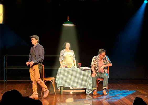 Cena de uma peça teatral em palco com fundo preto e iluminação cênica. À esquerda, um homem de boina, camisa escura e calça marrom caminha. Ao centro, uma mulher está em pé atrás de uma mesa coberta por toalha xadrez, sob um foco de luz. À direita, um homem sentado em uma cadeira olha para a mesa. Sobre ela, há objetos e uma taça. O piso é de madeira e a plateia aparece em silhueta na parte inferior.